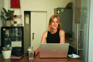 Een blonde vrouw zit in een koffiebar met haar laptop voor zich.